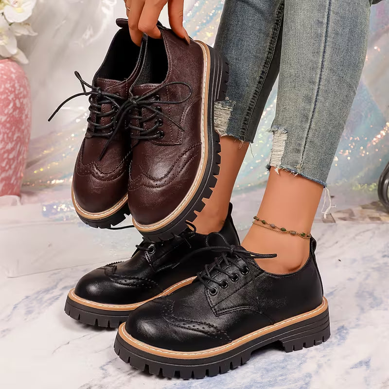 A person wearing a pair of black leather oxford-style platform shoes with brogue detailing and black laces, while holding a second pair of dark brown leather oxford shoes with the same sole design. The person is wearing distressed light-wash jeans and a gold anklet, standing on a light marble-patterned surface.