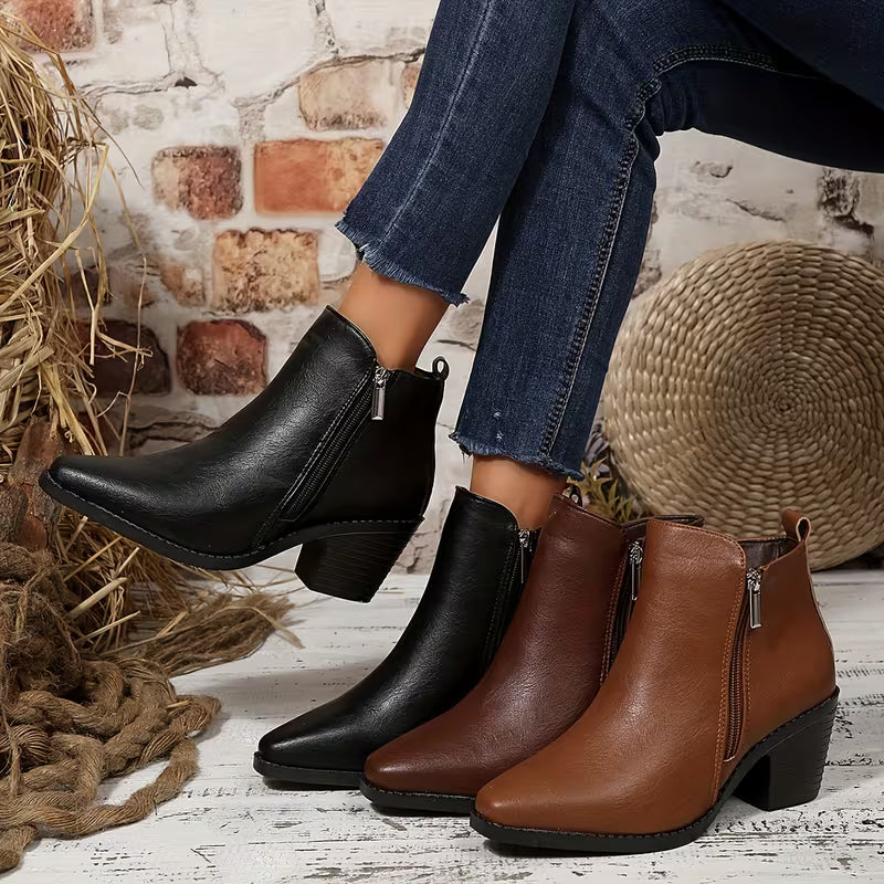 Three women's ankle boots with side zippers and block heels are shown on a rustic white wood floor with a brick wall backdrop. One boot is worn on a raised leg in blue jeans, while the other two sit on the floor in black and brown leather.