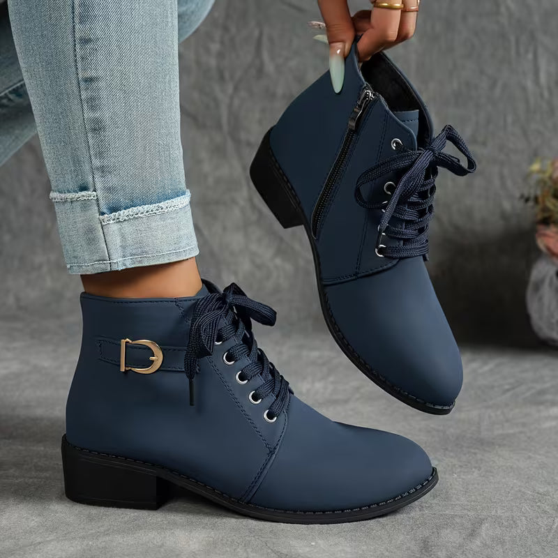 A pair of navy blue lace-up ankle boots with a side zipper, low block heel, and a gold buckle detail on the side, shown on a model wearing light-wash jeans while one boot is held up by a hand.