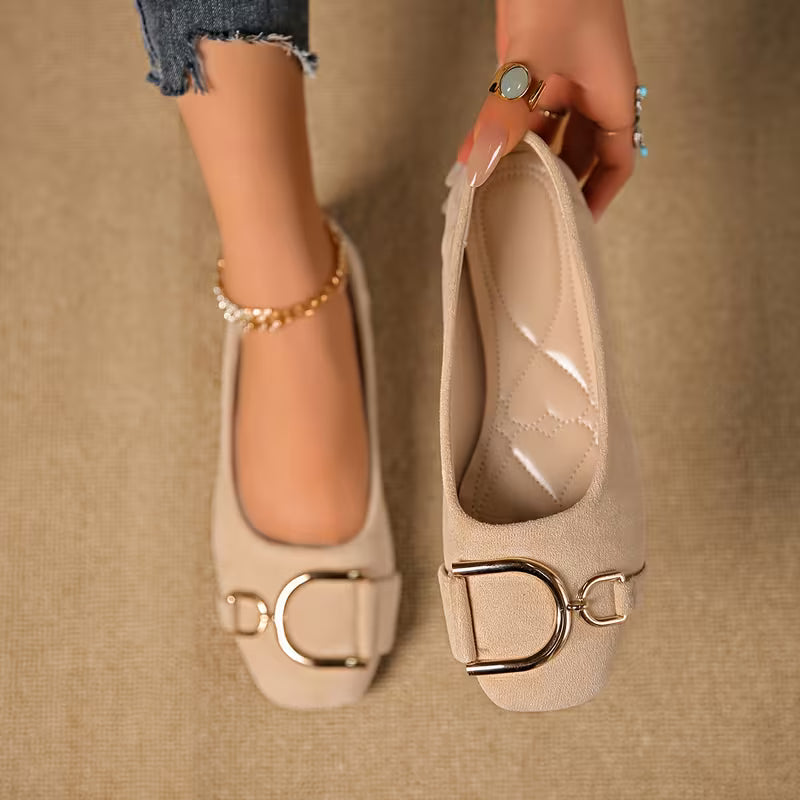 Beige flat shoes with gold buckles held by a hand on a beige background