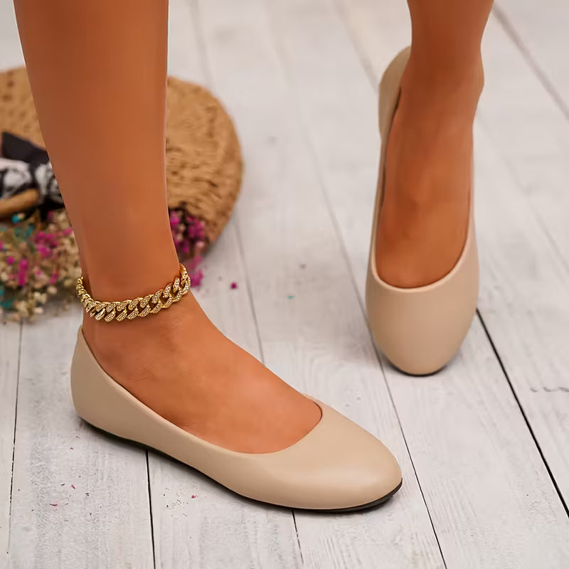 Beige flat shoes worn with a gold anklet on a light wooden floor.