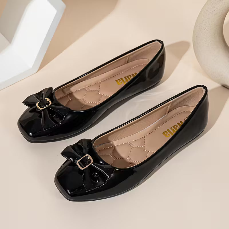 Black patent leather shoes with bow details on a beige surface