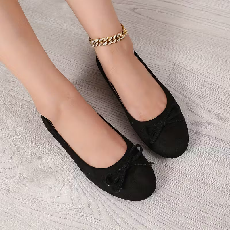 Black flats with bow detail worn by a person on a light wooden floor.
