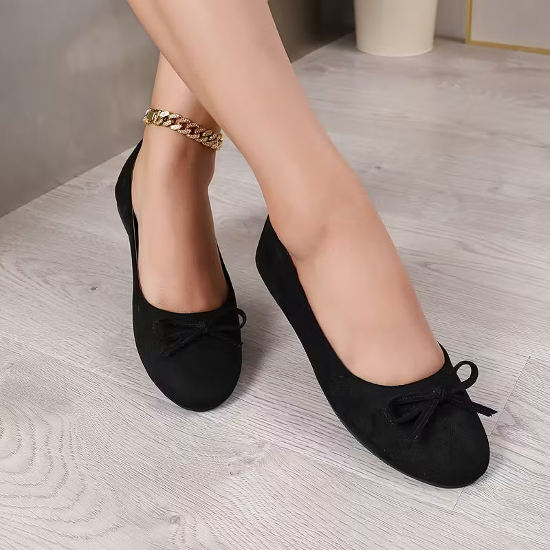 Black flat shoes with bows worn by a person on a light wooden floor.