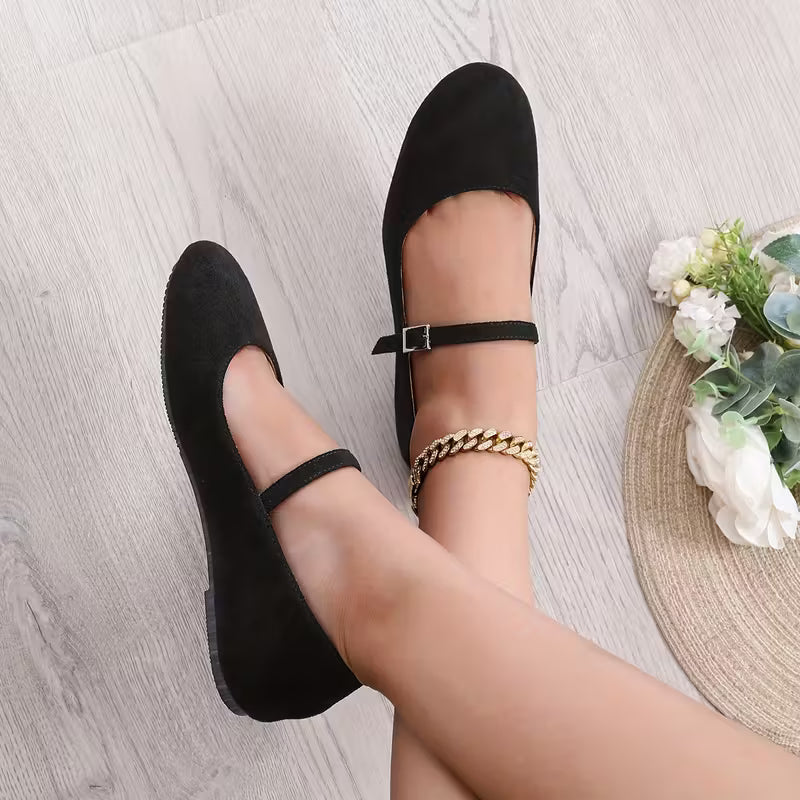 Black flat shoes worn on a light wooden floor with a decorative mat and flowers in the background.