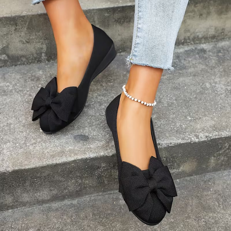 Black flat shoes with bow details worn on a stone step.