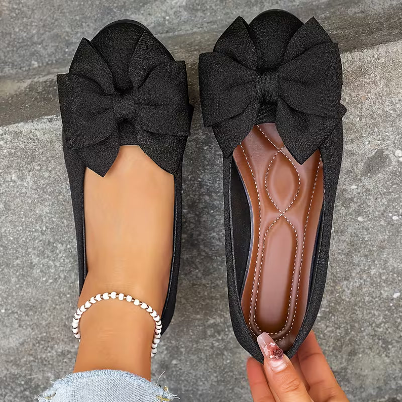 Black flat shoes with large bows worn by a person on a gray pavement background