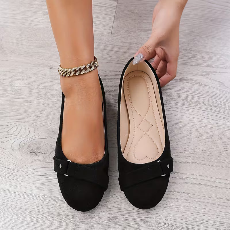 Black flat shoes with a bow held by a hand on a light wooden floor.