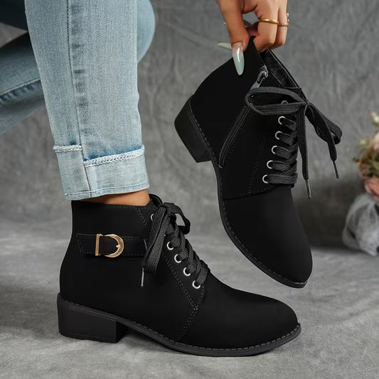 A person wearing black lace-up ankle boots with a side buckle strap, holding the other boot above their foot against a gray textured background.