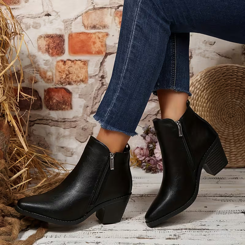 Black leather ankle boots with a pointed toe and block heel, shown on a model wearing blue jeans in a rustic setting with a brick wall, straw, and woven basket.