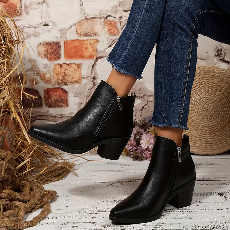 A person wearing black leather ankle boots with a block heel and side zipper, paired with blue jeans, posed in a rustic setting with a brick wall, straw, and woven basket.