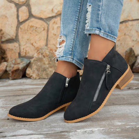 Black suede ankle boots with side zippers and stacked block heels, shown on a model wearing distressed blue jeans against a rustic stone wall and wooden floor.
