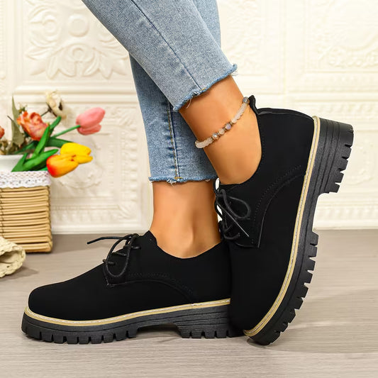 A pair of women's black lace-up oxford-style shoes with chunky lug soles and a thin beige stripe around the midsole, shown on a model wearing light blue jeans in a styled indoor setting.