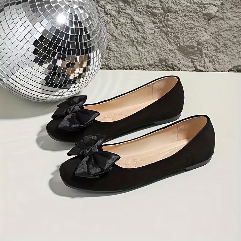 Black flats with bow details next to a disco ball on a light surface.