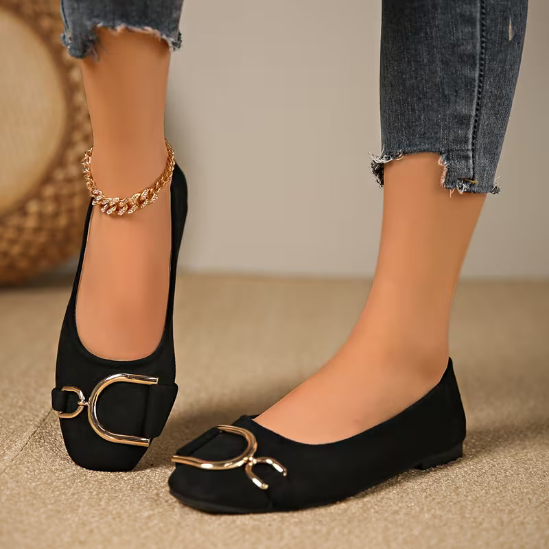 Black flats with gold buckles worn by a person on a neutral background
