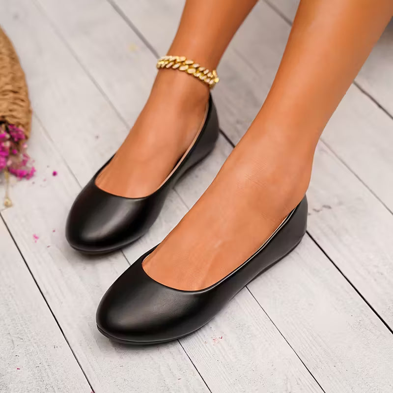 Black flat shoes worn on a light wooden floor with a gold anklet.