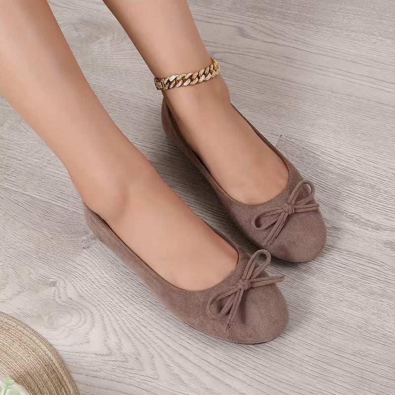Brown ballerina shoes with bows worn on a light wooden floor.