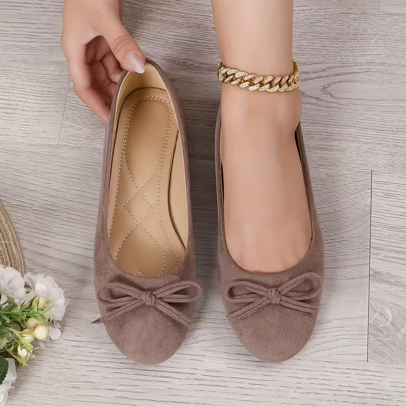 Brown suede flats with bow detail worn by a person on a wooden floor.