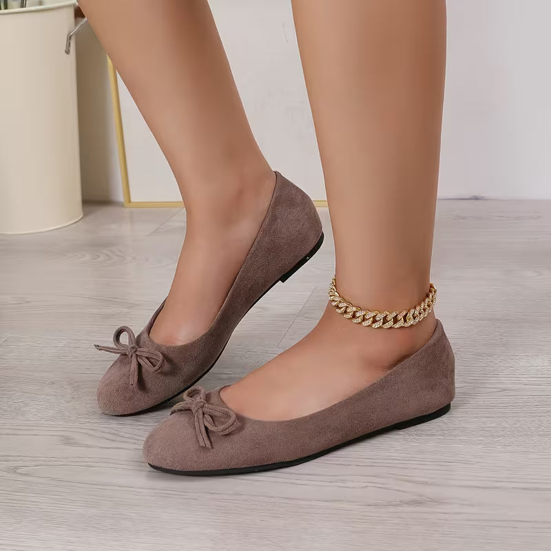 Brown suede shoes with bow details worn by a person on a light wooden floor.