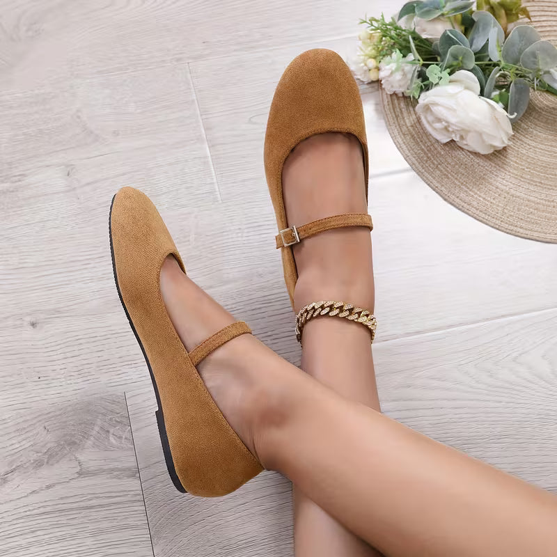 Brown suede shoes worn by a person on a light wooden floor with a decorative hat and flowers in the background.