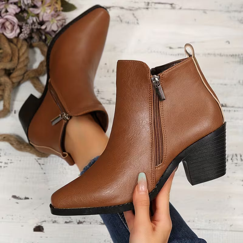 A pair of women's brown faux-leather ankle boots with a side zipper and block heel, shown on a foot and held by a hand over a rustic white wood background with flowers.