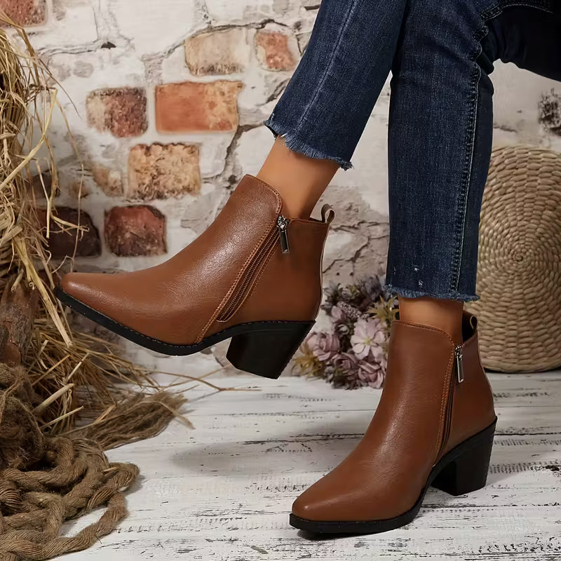 A pair of women's brown faux-leather ankle boots with a side zipper and block heel, shown on a model wearing blue jeans in a rustic setting with a brick wall, straw, and white wood floor.