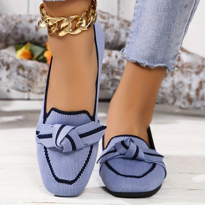 Blue loafers with a bow detail worn by a person, with a blurred background.