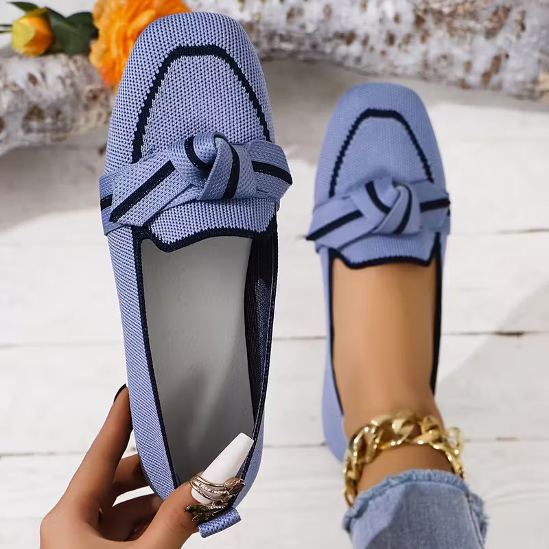 Blue loafers with a bow detail worn by a person on a light wooden floor.