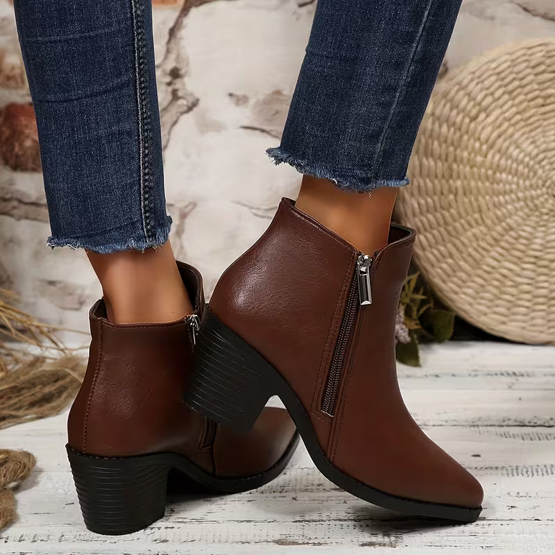 A pair of women's brown faux-leather ankle boots with black block heels and side zippers, shown on a model wearing blue jeans in a rustic styled setting.