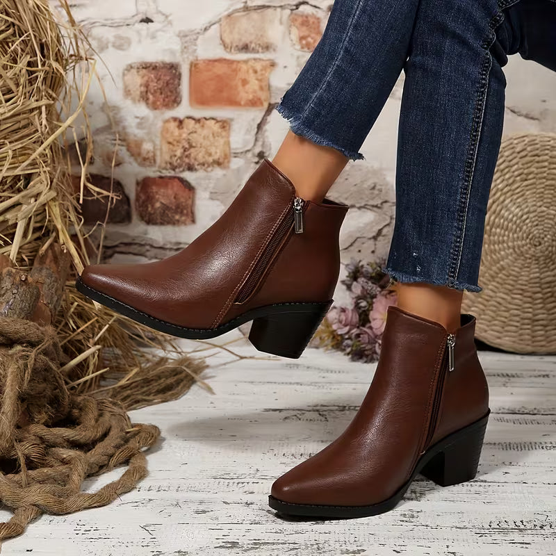 A person wearing brown leather ankle boots with side zippers and black block heels, paired with blue jeans, standing on a rustic white wooden floor with straw and rope decor.