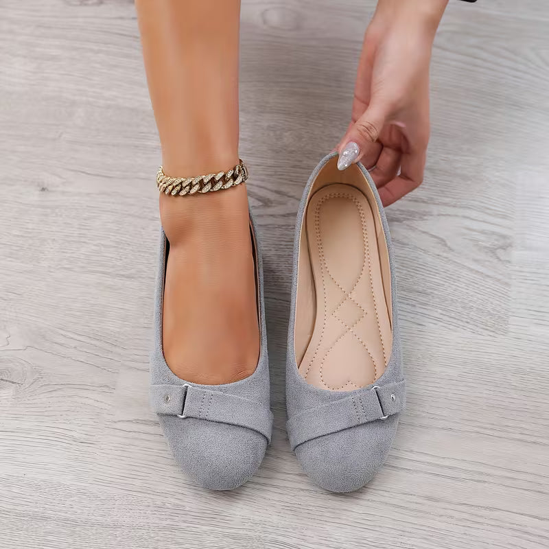 Gray suede shoes with a buckle held by a hand on a light wooden floor.