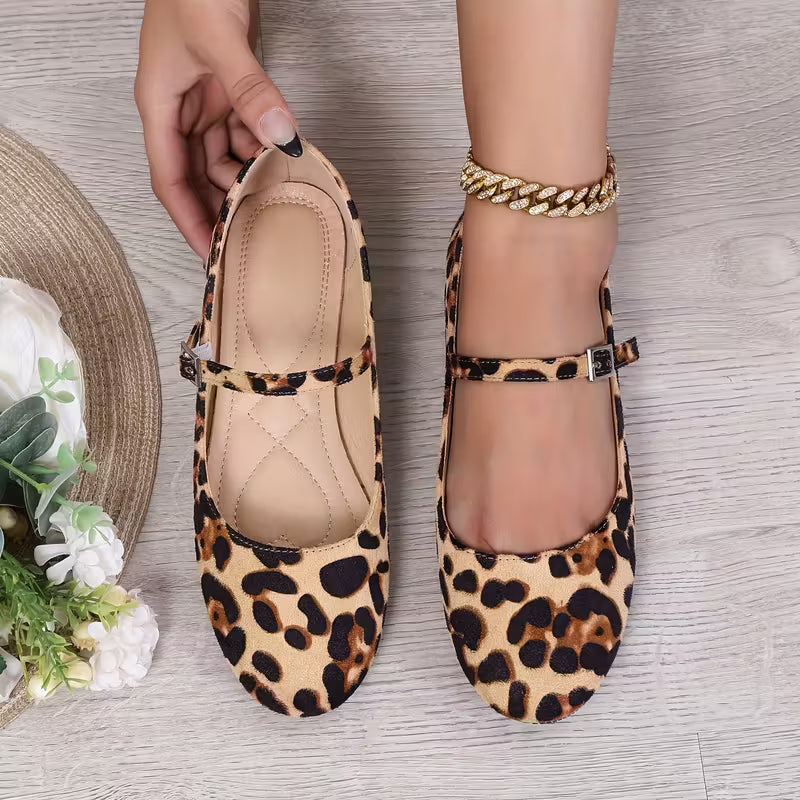 Leopard print shoes being held by a hand on a wooden surface with flowers and a hat.