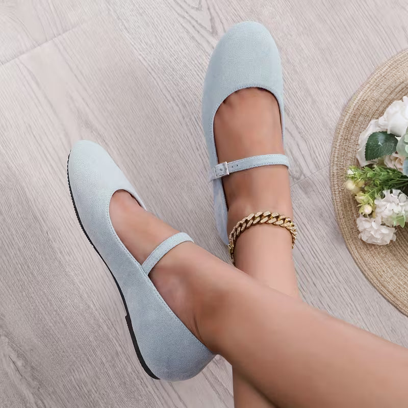 Light blue flats worn by a person on a light wooden floor with a woven mat and flowers in the background.