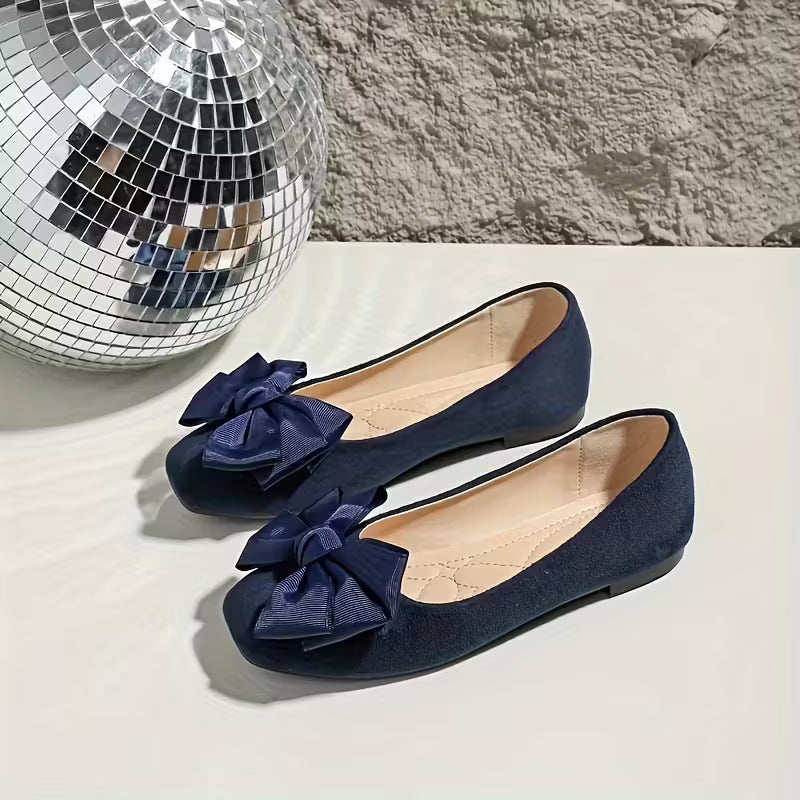 Navy blue shoes with large bows on a light surface next to a disco ball.