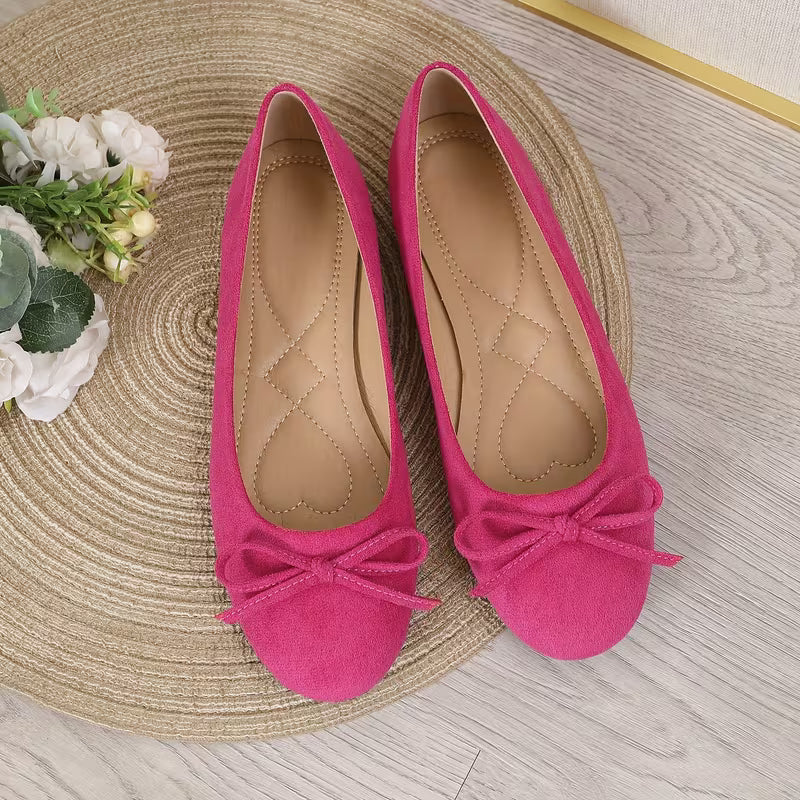Pair of pink suede shoes with bows on a woven mat with flowers.