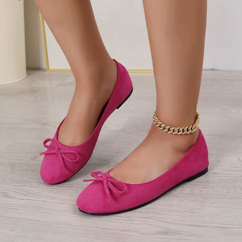 Pink suede shoes with bows worn by a person on a light wooden floor.