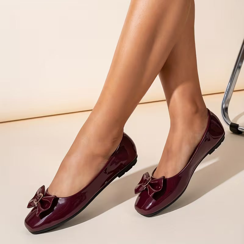 Burgundy patent leather flats with bow detail worn by a person on a light background