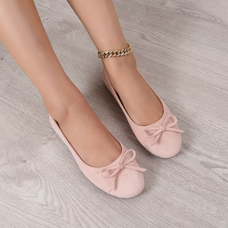Pink ballerina shoes with bows worn on a light wooden floor.