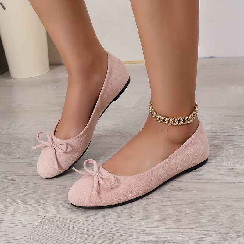 Pink suede shoes with bows worn by a person on a light wooden floor.
