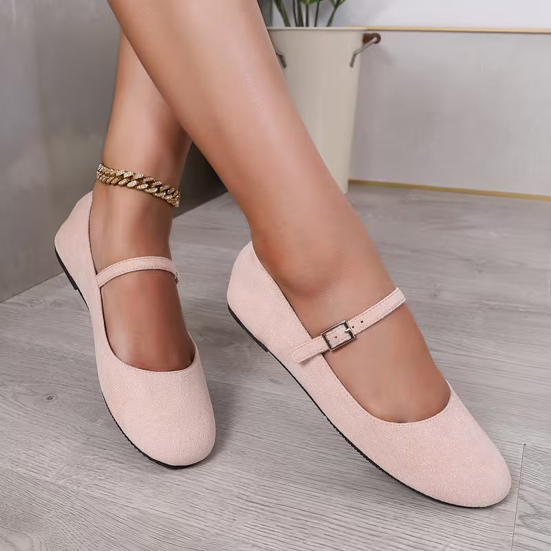Pink suede shoes with a strap worn by a person on a light wooden floor.