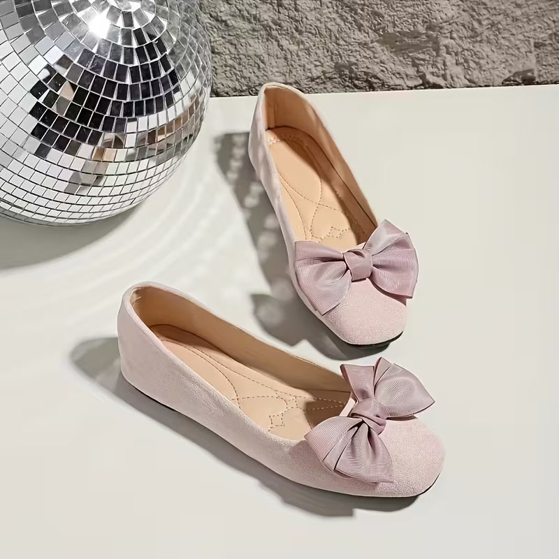 Pink flats with bow details on a light surface next to a disco ball.
