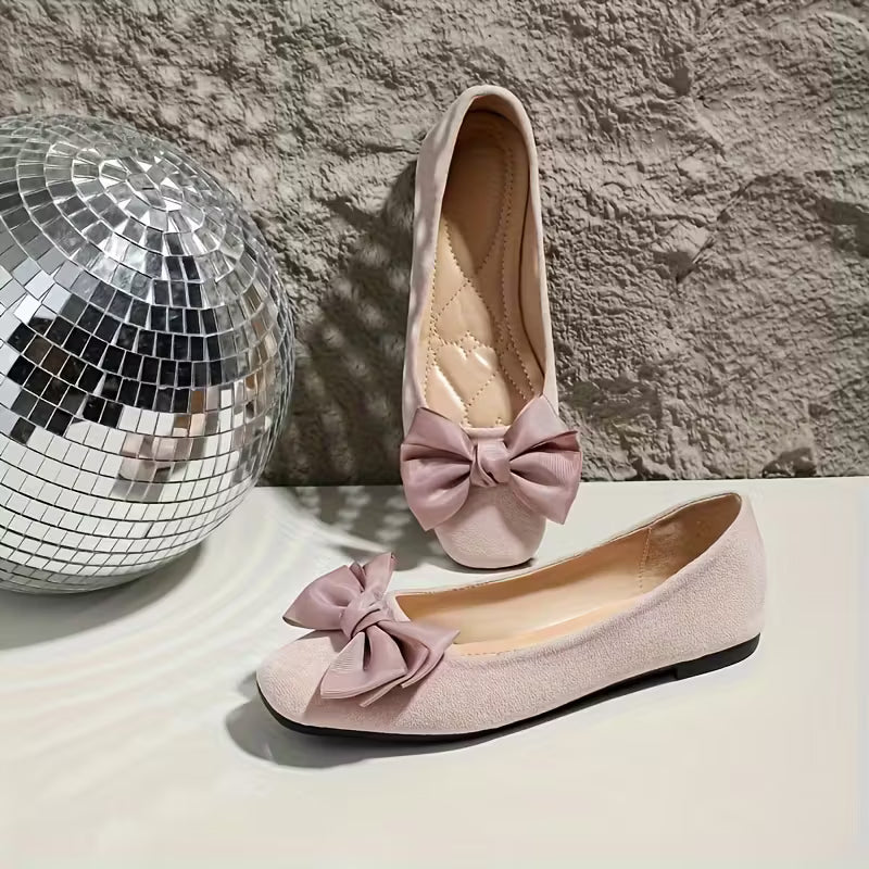 Pink ballet flats with bow details on a light surface next to a disco ball.