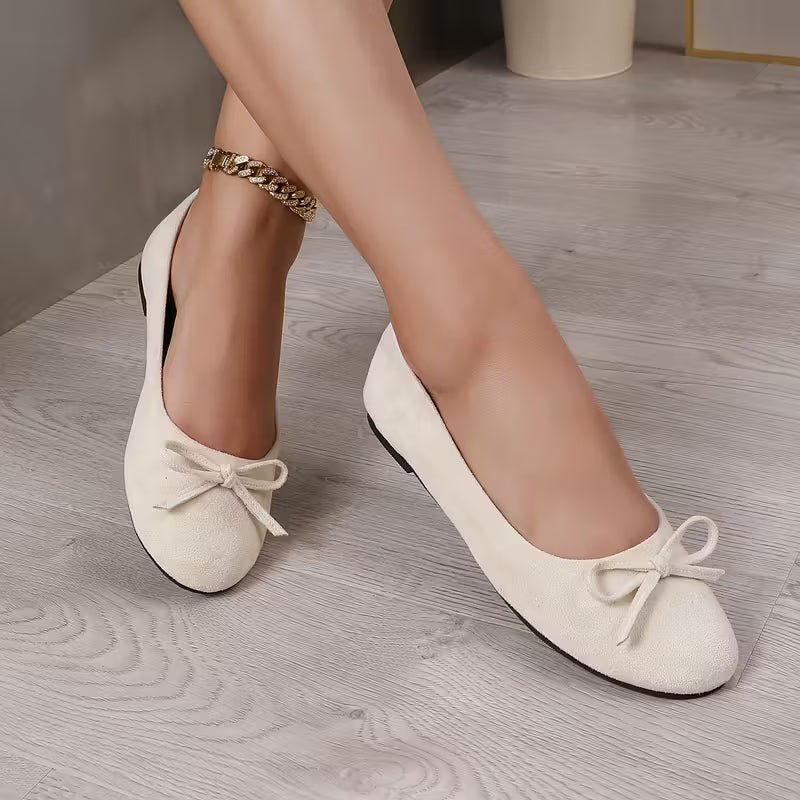 White ballerina shoes with bows worn by a person on a wooden floor.