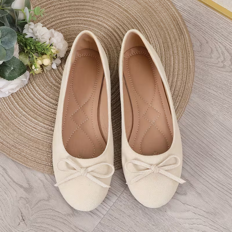 Pair of cream-colored flats with bow details on a textured surface.