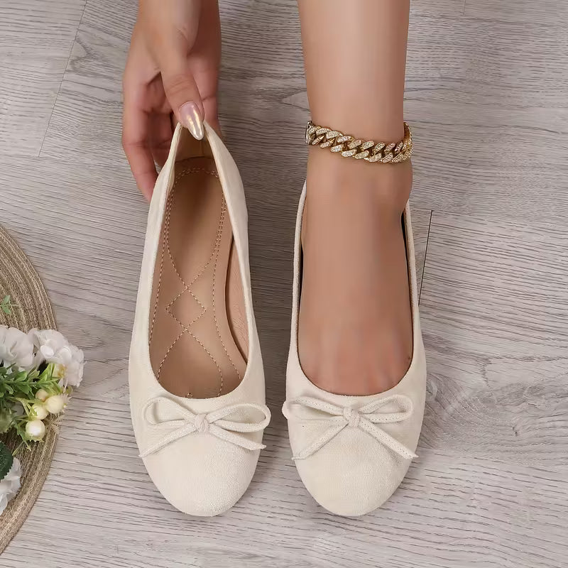 Beige flat shoes with bow details held by a hand on a wooden floor.