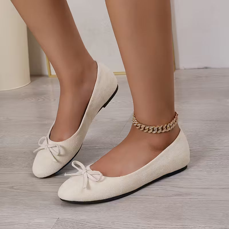 White flat shoes with black soles worn by a person on a light wooden floor.