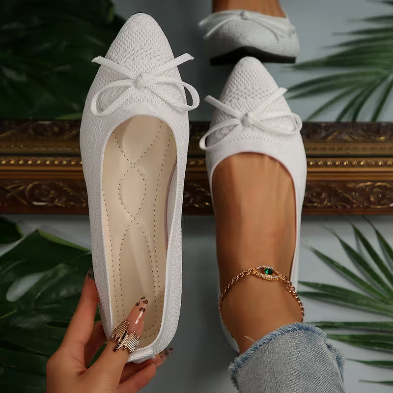 White pointed-toe shoes with a bow held by a hand, with a decorative mirror and green leaves in the background.