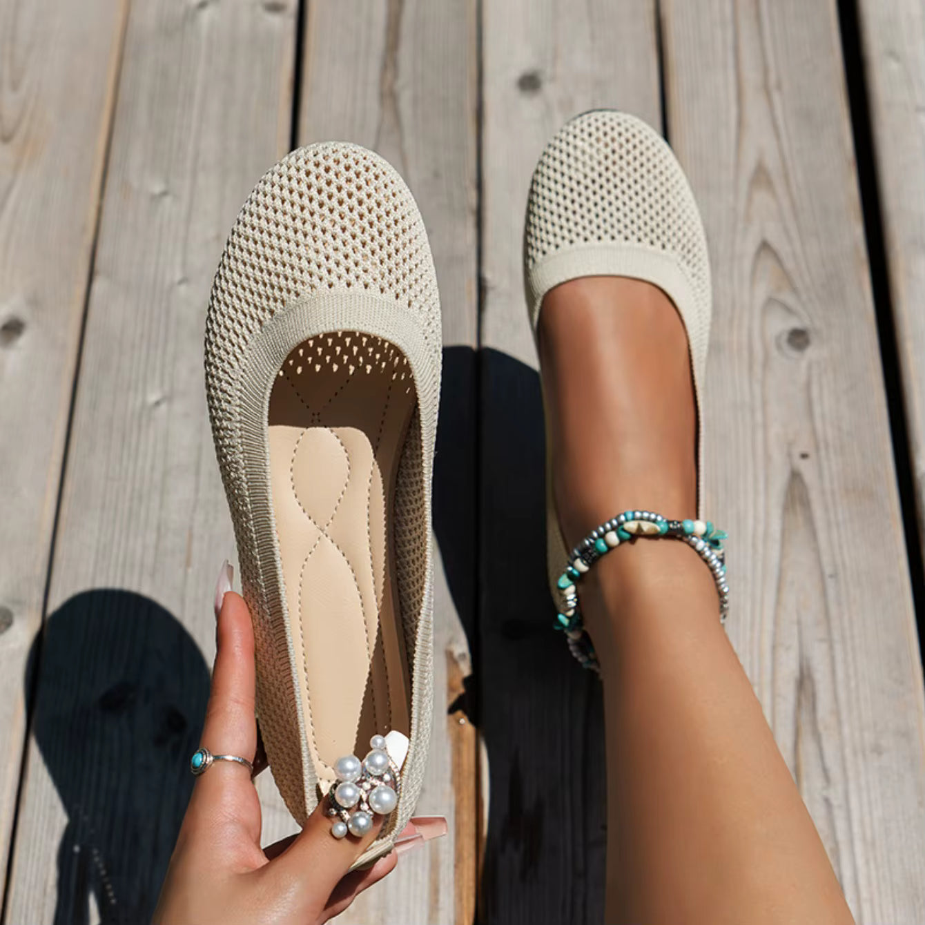Beige flats worn on a wooden floor with a hand holding one flat shoe.